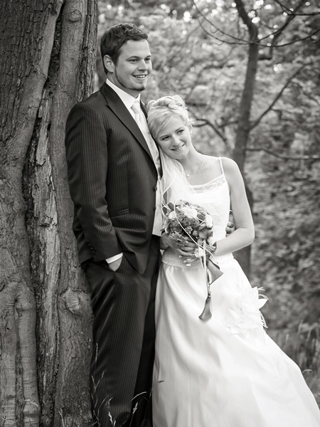 Hochzeitsportraet_Brautpaar_Baum_Schwarz-Weiss_Michael-Schalansky Schwarz-Weiss-Hochzeitsporträt eines lächelnden Brautpaares, das sich zärtlich an einen dicken Baumstamm in einem Wald oder Park lehnt. Die Braut trägt ein trägerloses Hochzeitskleid und hält einen Blumenstrauß. Der Bräutigam im Anzug hat eine Hand in der Hosentasche. Eine natürliche und romantische Hochzeitsfotografie von Michael Schalansky.